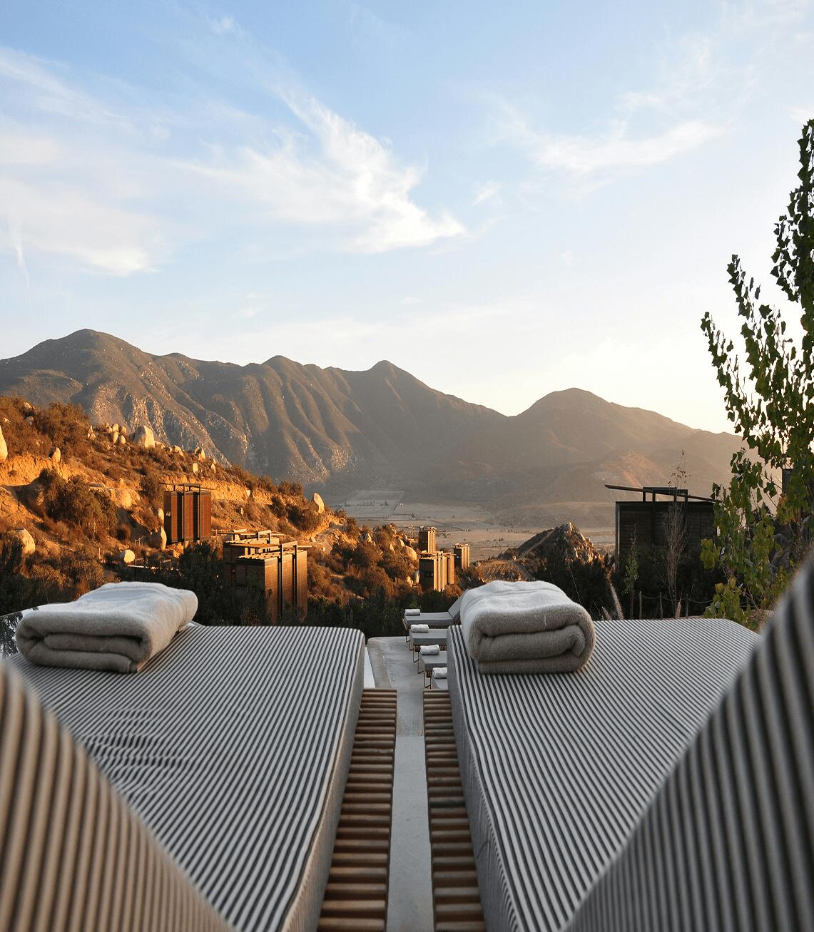 Duo of lounge chairs topped with neatly folded blankets overlooking a stunning valley view of a rugged mountainous landscape in late afternoon. 