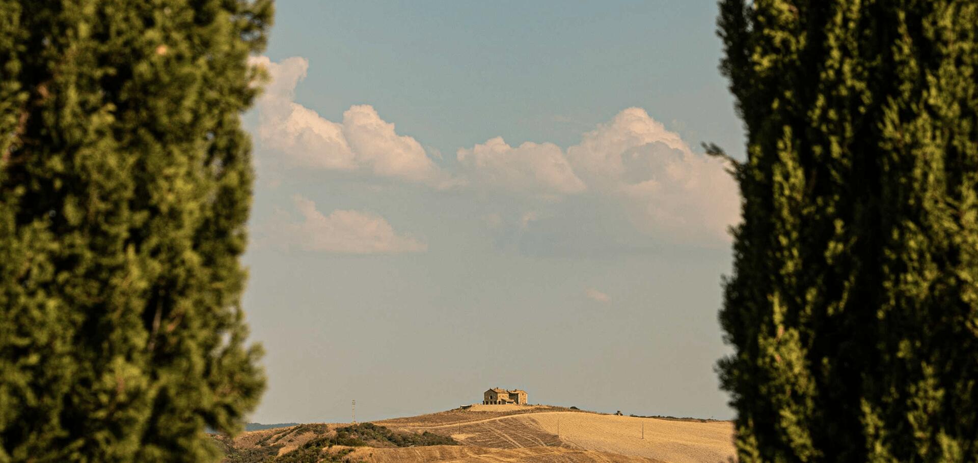 Tuscan landscape featuring a solitary farmhouse perched on a hill framed by two cypresses.