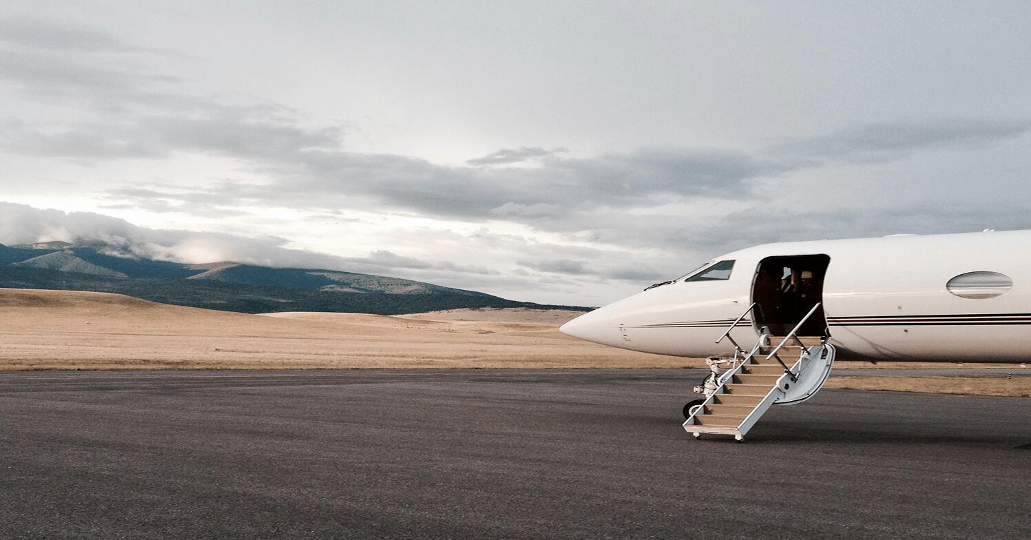 A private gulfstream jet sits on a remote airstrip, its stairs lowered against a dramatic backdrop of rolling golden plains and mountains.